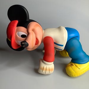 May include: A vintage Mickey Mouse figure in a crawling position. The figure has a black head with red details, a white and blue outfit, red arms, and yellow shoes. The toy is made of a hard plastic material.