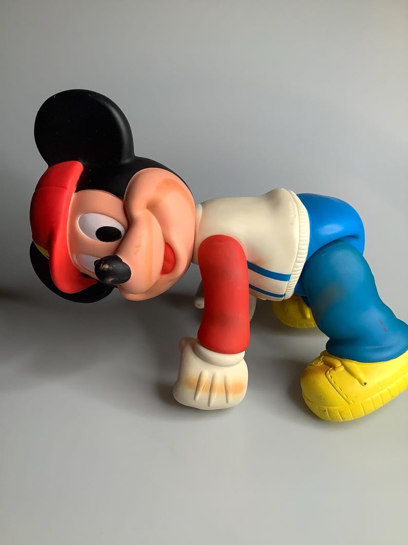 May include: A vintage Mickey Mouse figure in a crawling position. The figure has a black head with red details, a white and blue outfit, red arms, and yellow shoes. The toy is made of a hard plastic material.