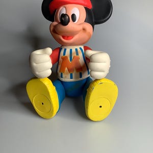 May include: A vintage Mickey Mouse figurine. The figure is wearing a red cap, a white and blue striped shirt with an orange "M", blue shorts, and yellow shoes. The figure has black ears, white gloves, and a smiling face.