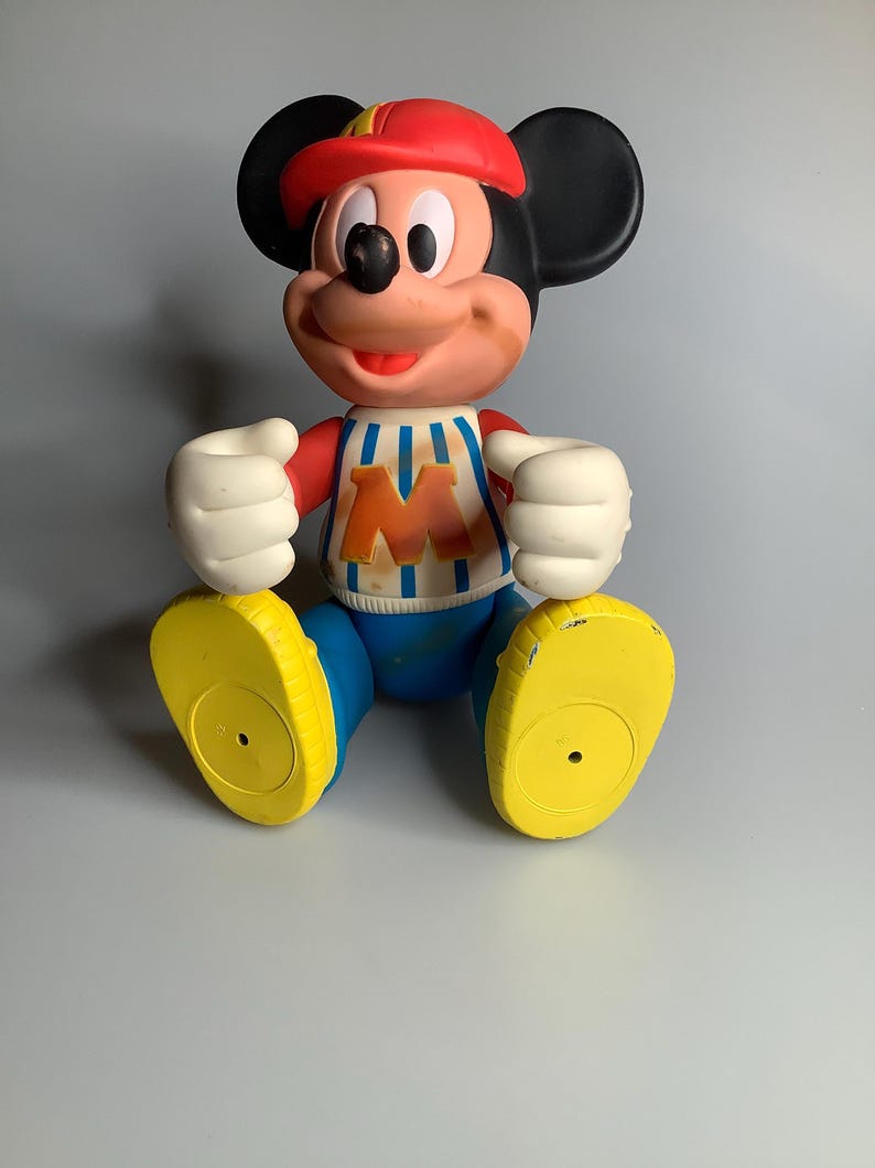 May include: A vintage Mickey Mouse figurine. The figure is wearing a red cap, a white and blue striped shirt with an orange "M", blue shorts, and yellow shoes. The figure has black ears, white gloves, and a smiling face.