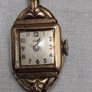 May include: A vintage gold-tone wristwatch with a square face, featuring a white dial with black Arabic numerals and a small crown on the right side. The case has decorative metal lugs at the top and bottom, connected to a metallic link band.