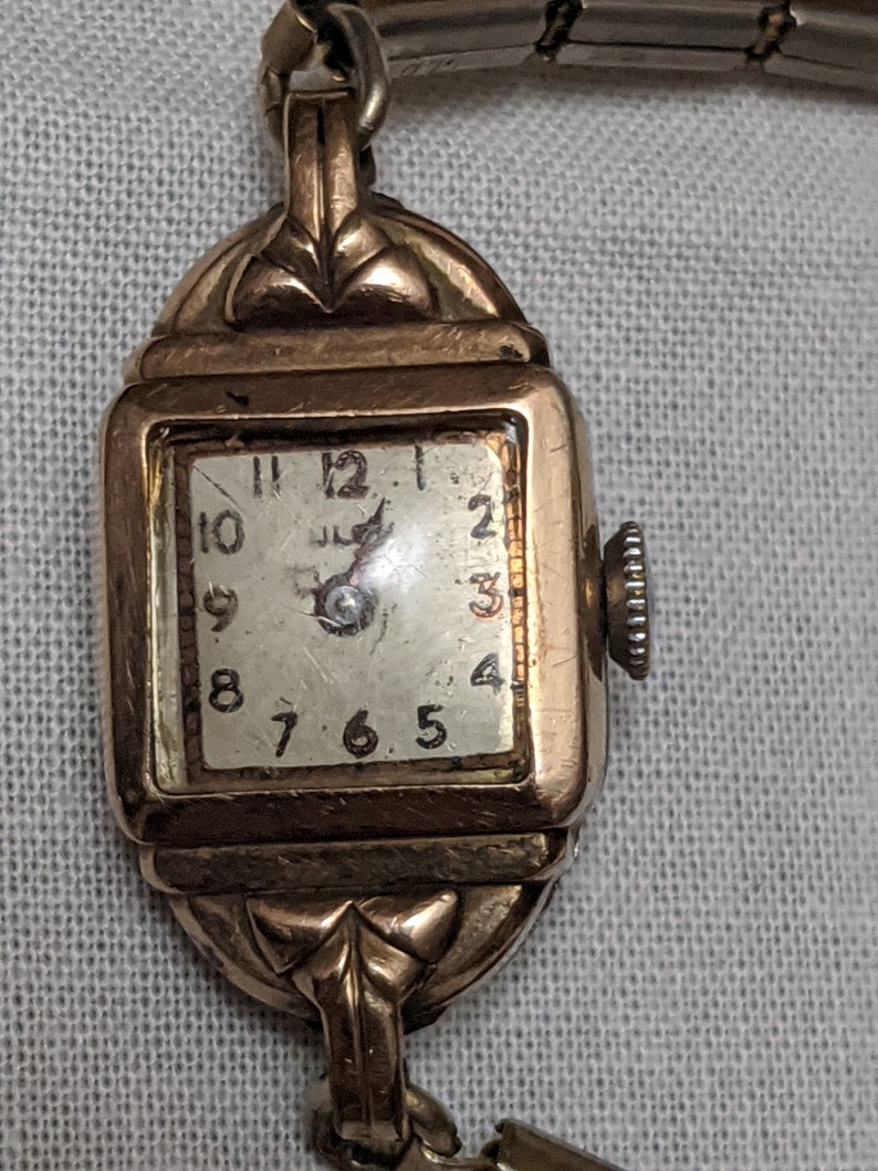 May include: A vintage gold-tone wristwatch with a square face, featuring a white dial with black Arabic numerals and a small crown on the right side. The case has decorative metal lugs at the top and bottom, connected to a metallic link band.
