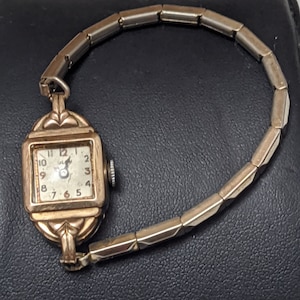May include: A vintage gold-tone wristwatch with a square face, featuring a white dial with black Arabic numerals and a small crown on the side. The watch has an expandable metal link bracelet with a polished finish and decorative lugs.