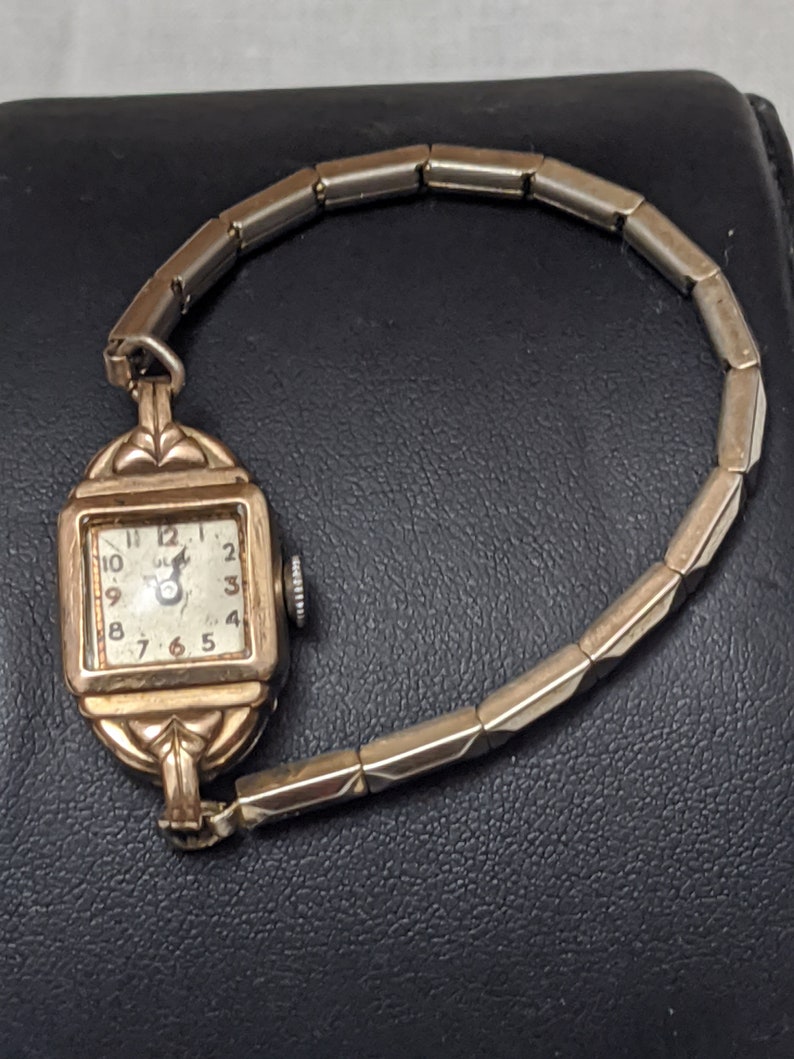 May include: A vintage gold-tone wristwatch with a square face, featuring a white dial with black Arabic numerals and a small crown on the side. The watch has an expandable metal link bracelet with a polished finish and decorative lugs.