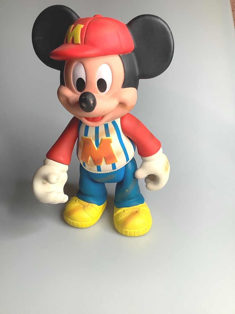 May include: A Mickey Mouse figurine wearing a red baseball cap with a yellow "M", a red and white striped shirt, blue trousers, and yellow shoes. The figure has black ears, white gloves, and a smiling expression.
