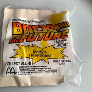 May include: A yellow and red McDonald's Happy Meal bag featuring the Back to the Future logo and Marty's Hoverboard. The bag says "Collect All 4!" and "While Supplies Last".
