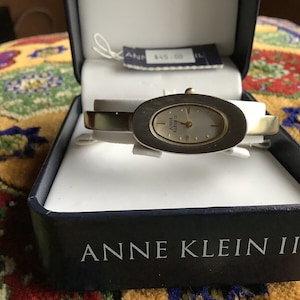 May include: A silver and gold oval-shaped wristwatch with a white face and black Roman numeral markings. The watch is in a blue box with the words "Anne Klein II" printed on the front.