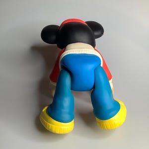 May include: A large Mickey Mouse figurine, viewed from the back. It features black ears, a red jacket, blue shorts, and yellow shoes. This colourful figurine is a collectible item.