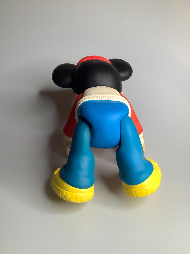 May include: A large Mickey Mouse figurine, viewed from the back. It features black ears, a red jacket, blue shorts, and yellow shoes. This colourful figurine is a collectible item.