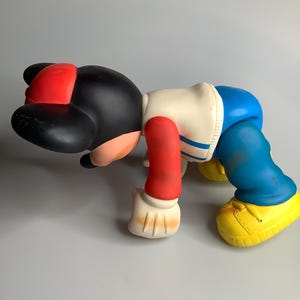 May include: A colourful vinyl figure in a crawling pose. The figure has a black head with a red cap, a white and blue jacket, red arms, blue trousers, and yellow shoes. The figure is a collectible toy.