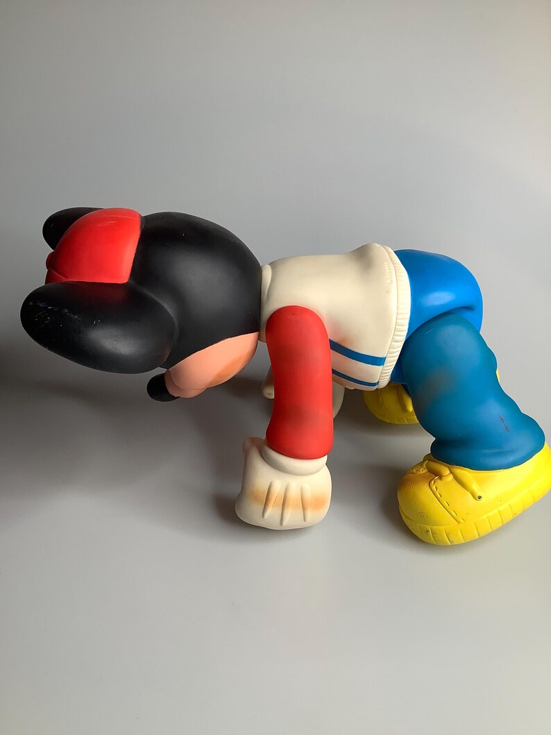 May include: A colourful vinyl figure in a crawling pose. The figure has a black head with a red cap, a white and blue jacket, red arms, blue trousers, and yellow shoes. The figure is a collectible toy.