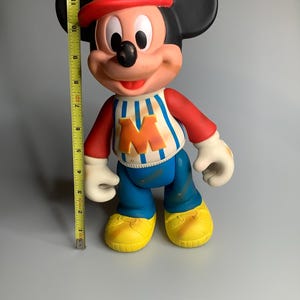 May include: A vintage Mickey Mouse figurine, approximately 28 cm tall, wearing a red cap, a blue and white striped shirt with an orange "M", blue trousers, and yellow shoes. The figure has black ears and white gloves.