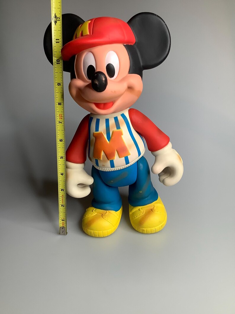 May include: A vintage Mickey Mouse figurine, approximately 28 cm tall, wearing a red cap, a blue and white striped shirt with an orange "M", blue trousers, and yellow shoes. The figure has black ears and white gloves.