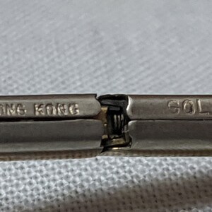 May include: A metallic hinge mechanism from a piece of jewelry or accessory. The left side is stamped with the text HONG KONG and the right side is stamped with the word GOLD. The metal has a silver-toned finish with a visible spring hinge.