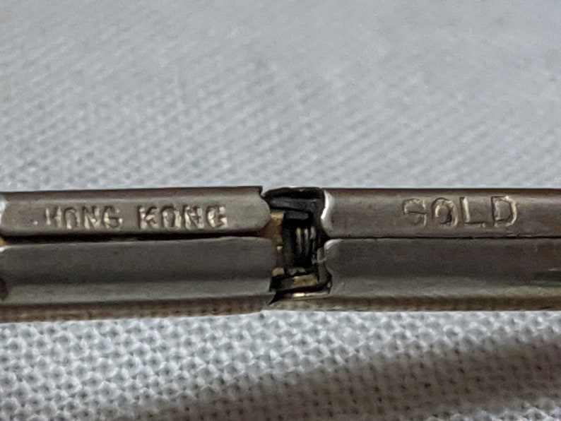 May include: A metallic hinge mechanism from a piece of jewelry or accessory. The left side is stamped with the text HONG KONG and the right side is stamped with the word GOLD. The metal has a silver-toned finish with a visible spring hinge.