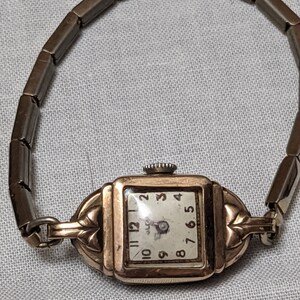 May include: A gold-tone square wristwatch with a white dial featuring black Arabic numerals and a matching flexible metal link band. The watch case has decorative leaf-like accents on the sides and a small winding crown at the top.