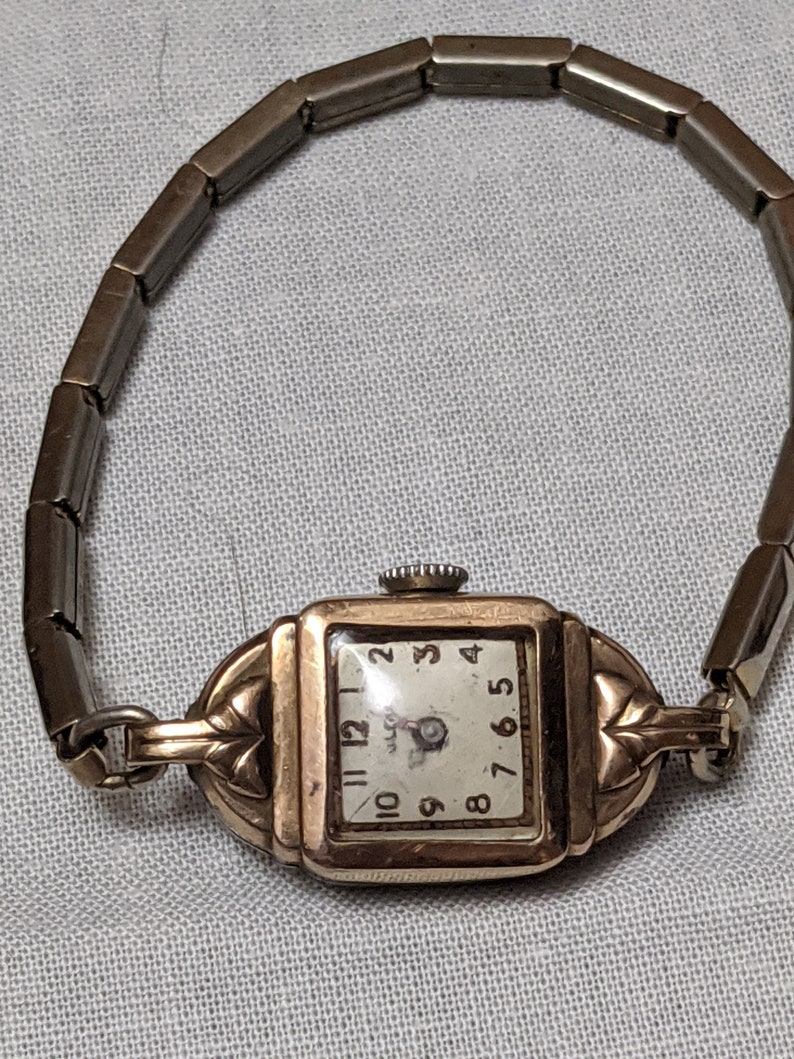 May include: A gold-tone square wristwatch with a white dial featuring black Arabic numerals and a matching flexible metal link band. The watch case has decorative leaf-like accents on the sides and a small winding crown at the top.