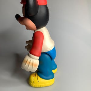 May include: A vintage Mickey Mouse figurine in profile. The figure is wearing a red cap, black ears, and a white shirt with red sleeves. Blue trousers and yellow shoes complete the outfit. The figurine is a classic cartoon character.