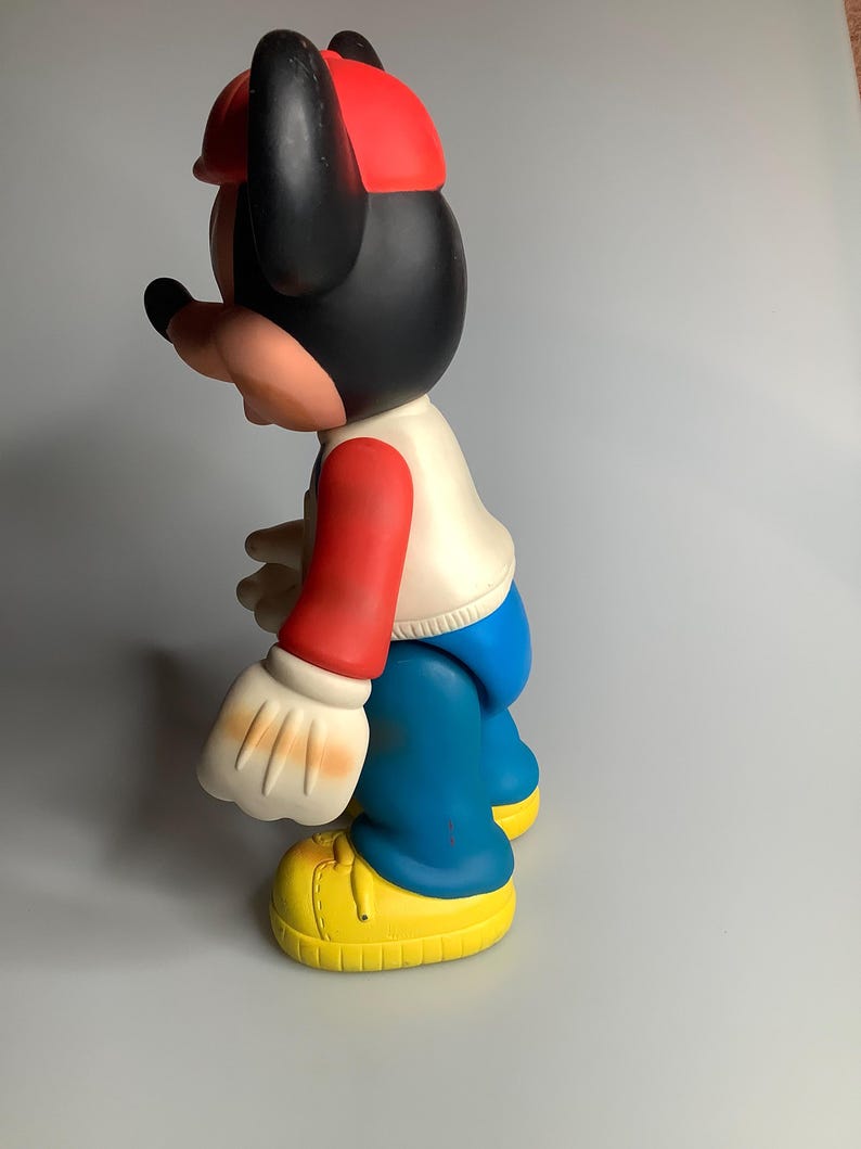 May include: A vintage Mickey Mouse figurine in profile. The figure is wearing a red cap, black ears, and a white shirt with red sleeves. Blue trousers and yellow shoes complete the outfit. The figurine is a classic cartoon character.