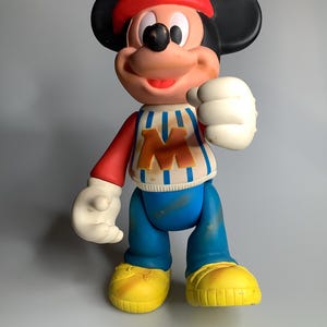 May include: A vintage Mickey Mouse figure. The figure is wearing a red cap, a striped top with a large "M", blue trousers, and yellow shoes. The figure has black ears, white gloves, and a smiling expression.