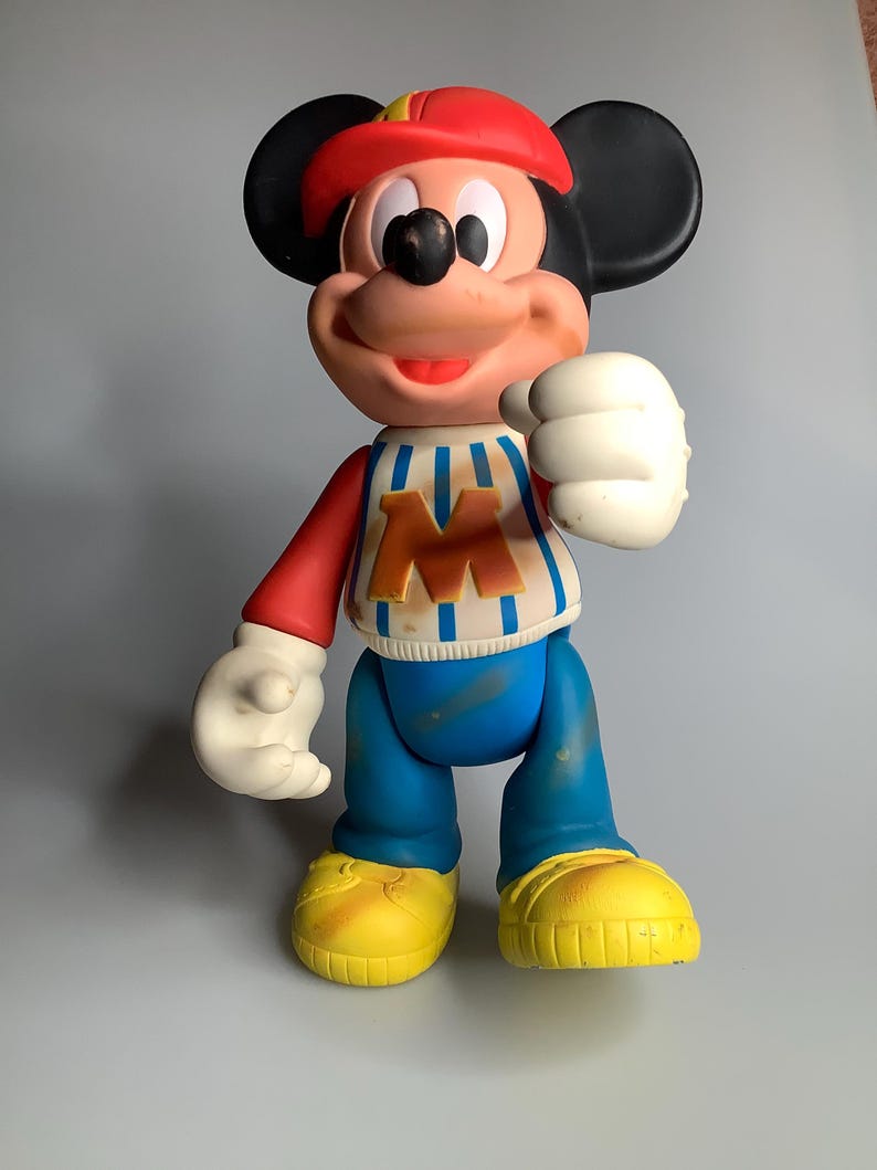 May include: A vintage Mickey Mouse figure. The figure is wearing a red cap, a striped top with a large "M", blue trousers, and yellow shoes. The figure has black ears, white gloves, and a smiling expression.