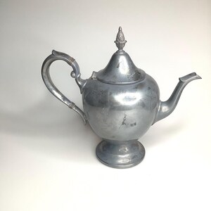 May include: A silver-toned metal teapot with a rounded body, a curved handle, and a pointed spout. The lid features a decorative finial at the top. The surface of the teapot shows signs of wear and oxidation, giving it a matte, aged appearance.