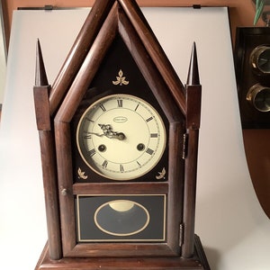 May include: A dark brown wooden clock with a steeple design. The clock face has a white background with black Roman numerals and black hands. The clock is made by the company "Ethan Allen".