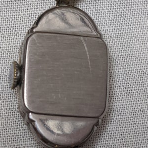 May include: A vintage-style wristwatch featuring a rectangular silver-toned case with rounded edges and a textured winding crown on the side. The watch is attached to a metallic link bracelet, showcasing a polished finish and visible light surface scratches.