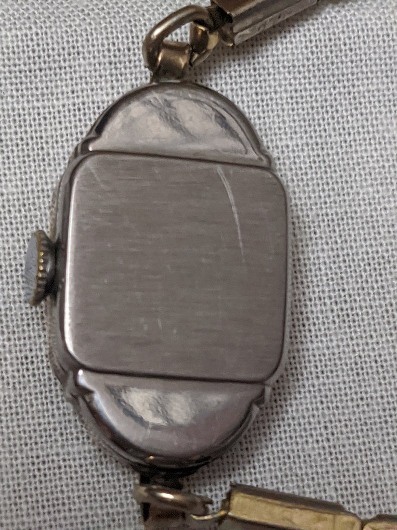 May include: A vintage-style wristwatch featuring a rectangular silver-toned case with rounded edges and a textured winding crown on the side. The watch is attached to a metallic link bracelet, showcasing a polished finish and visible light surface scratches.