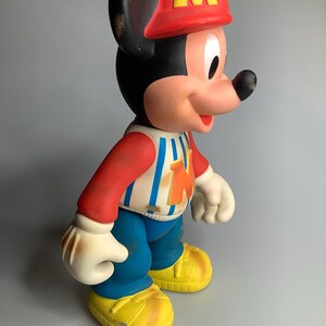 May include: A vintage Mickey Mouse figure, wearing a red hat with a yellow "M", a red and white striped shirt, blue trousers, and yellow shoes. The classic cartoon character is in a standing pose.