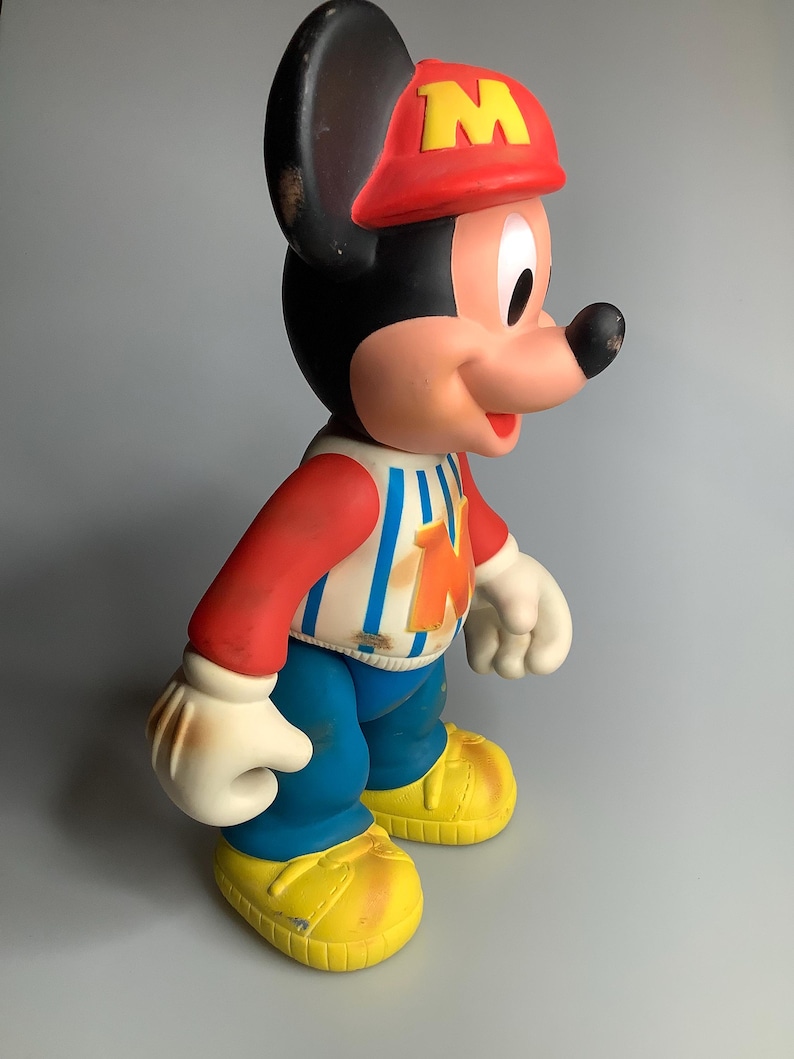 May include: A vintage Mickey Mouse figure, wearing a red hat with a yellow "M", a red and white striped shirt, blue trousers, and yellow shoes. The classic cartoon character is in a standing pose.
