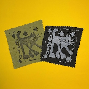 May include: Two square fabric patches with pinked edges. One patch is olive green with a dark brown cat illustration and the word GATO. The other patch is black with a silver cat illustration and the word GATO. Both feature star accents.