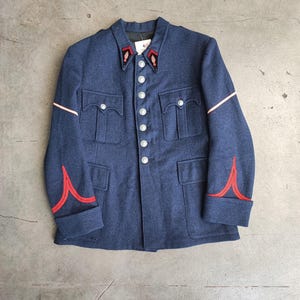Peut inclure: Veste militaire bleu marine vintage avec des boutons argentés et quatre poches avant. La veste présente des bordures rouges et blanches sur le col, les manches et les poignets. La veste est en matière de type laine.