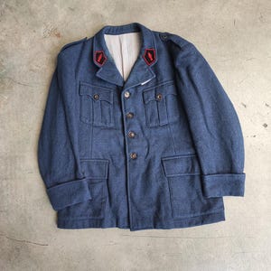 Peut inclure: Veste en laine bleue avec quatre poches avant, des boutons et des détails rouges et noirs sur les épaules. La veste a un col et des poignets repliés. Style vintage.