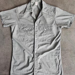 May include: A pale grey short-sleeved shirt with two chest pockets and a button-up front. The shirt has a collar and is labelled "1930/40's FRENCH LINEN/COTTON OLD FASHION SHIRT" and "SMALL".