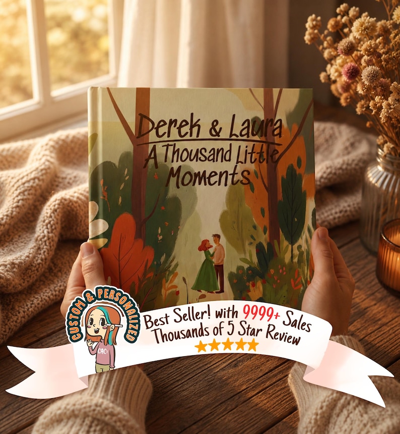 May include: A personalized book cover with the title "Derek & Laura: A Thousand Little Moments." The cover features an illustration of a couple in a forest setting with autumn colors. The book is held by a person.