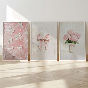 以下が含まれることがあります： 3つの額入りアートプリント。最初のものは、ピンクの背景に白いリボンが描かれています。2つ目は、テクスチャ加工された白い背景にピンクのリボンがあります。3つ目は、花瓶に入ったピンクのアジサイを描いています。すべてニュートラルなフレームに入っています。