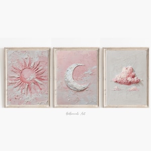 May include: Three framed wall art pieces featuring celestial themes. The first depicts a pink sun with textured rays, the second a white crescent moon, and the third a pink cloud. The artwork is set against a soft pink and white background.