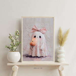 May include: A framed artwork featuring a whimsical ghost character. The ghost is white with a pink bow and cheeks, holding a small pumpkin with a lit candle. The artwork has a textured, painterly style, with a soft color palette of pinks, whites, and oranges. The artwork is displayed on a wooden shelf.