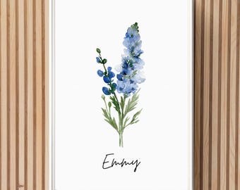 Könnte beinhalten: Ein Aquarellbild einer blauen Blume mit grünen Stielen und Blättern. Das Bild ist in einem weißen Rahmen gerahmt und der Name "Emmy" ist in schwarzer Kursivschrift unter der Blume geschrieben.