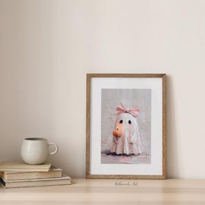 May include: A framed art print featuring a cute ghost illustration. The ghost is depicted in soft pink hues, wearing a bow and holding a small pumpkin. The artwork has a textured, painterly style, perfect for Halloween decor.
