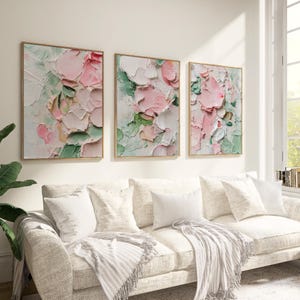 May include: Three framed floral paintings in a textured, impasto style. The artwork features pink and white flowers with green leaves against a light background. The frames are a light gold colour. The paintings are displayed above a white sofa with pillows.