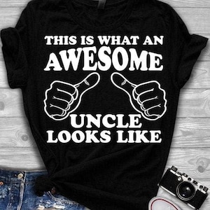 May include: Black t-shirt with white text that reads "THIS IS WHAT AN AWESOME UNCLE LOOKS LIKE." The design includes two thumbs-up hand gestures. The shirt is folded, and a camera and part of a pair of jeans are visible.