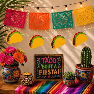 May include: A festive party decoration set featuring a string of colorful papel picado banners, a string of taco-shaped cutouts, and a wooden sign with the text TACO 'BOUT A FIESTA! in pink, green, and yellow letters, accented by floral pots.