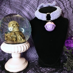 May include: A purple fuzzy choker with a silver ring and a light purple bell charm. The choker is displayed on a black mannequin. A glass snow globe with a skull inside is in the foreground.