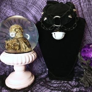May include: A black velvet choker with silver spikes and a silver chain with a white bell. The choker is displayed on a black mannequin bust. The bust is on a purple and black mesh background.