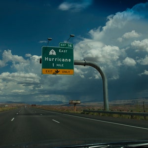 May include: A green highway sign with white text reads "Exit 16 East Hurricane 1 Mile Exit Only". The sign is mounted on a metal pole with a curved arm. The sign is visible against a blue sky with white clouds.