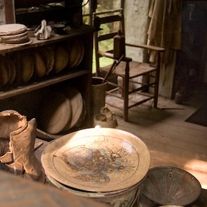 May include: A wooden shelf displaying a collection of pottery items, including plates, bowls, and figurines. The shelf is in a rustic setting with a wooden floor and a brick wall in the background.