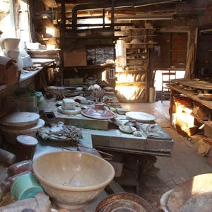 May include: A cluttered workshop with wooden shelves and tables filled with pottery and ceramic pieces. There are various sizes and shapes of pottery, including bowls, plates, and vases. The workshop is lit by natural light coming in from a window.