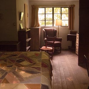 May include: A bedroom with a patchwork quilt on the bed, a wooden dresser with a lamp on top, and a window with a view of trees. There is a chair in the corner of the room.
